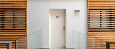 Entrada de un edificio moderno con una puerta blanca marcada “209”, paneles de madera con listones en las paredes y una barandilla de cristal al frente.