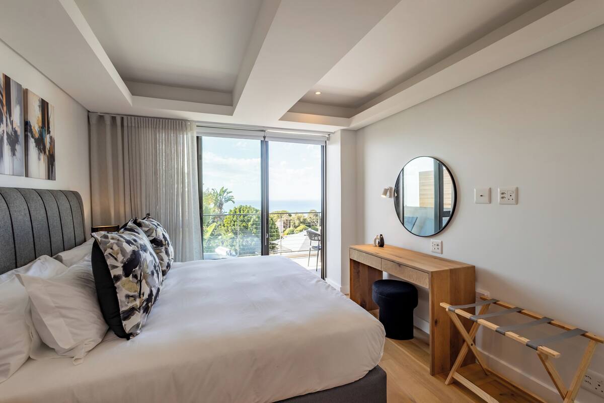 A modern hotel room with a large bed, a wooden desk, a round mirror, and a balcony with a view.