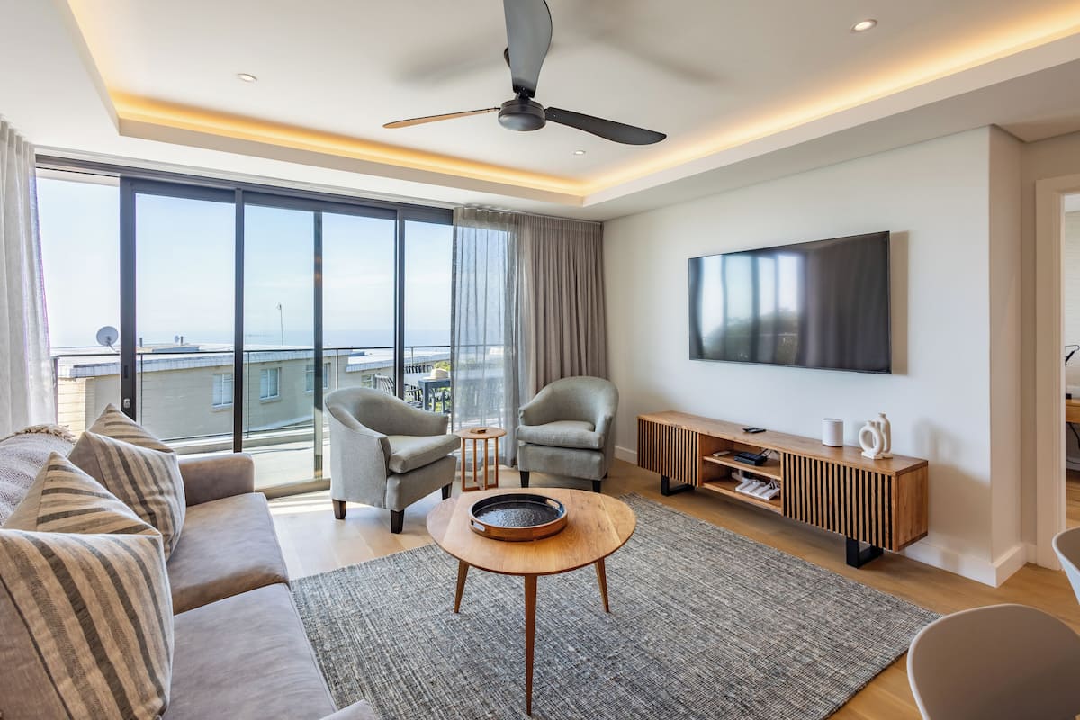 A modern living room with a large flat-screen TV, a ceiling fan, and a view of the city through large windows.