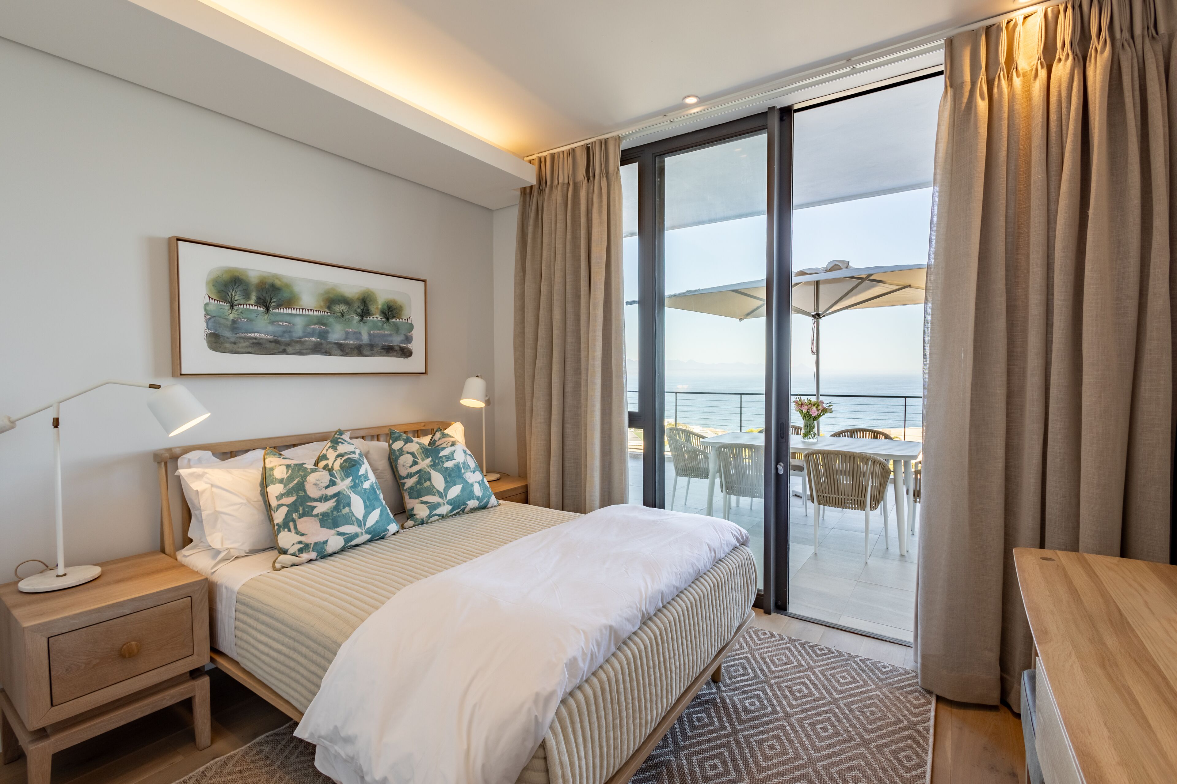 Habitación de hotel con cama, mesita de noche, lámpara y vistas a la terraza con mobiliario exterior.