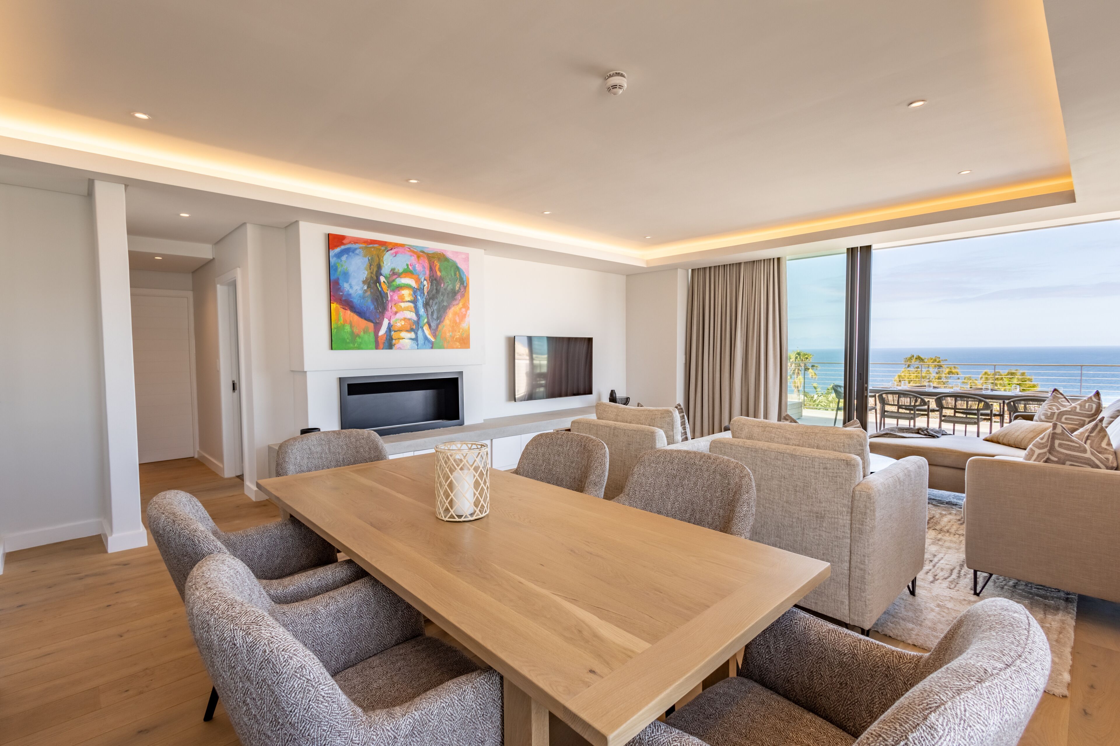A modern living room with a wooden dining table, comfortable seating, a flat-screen TV, and a large painting.
