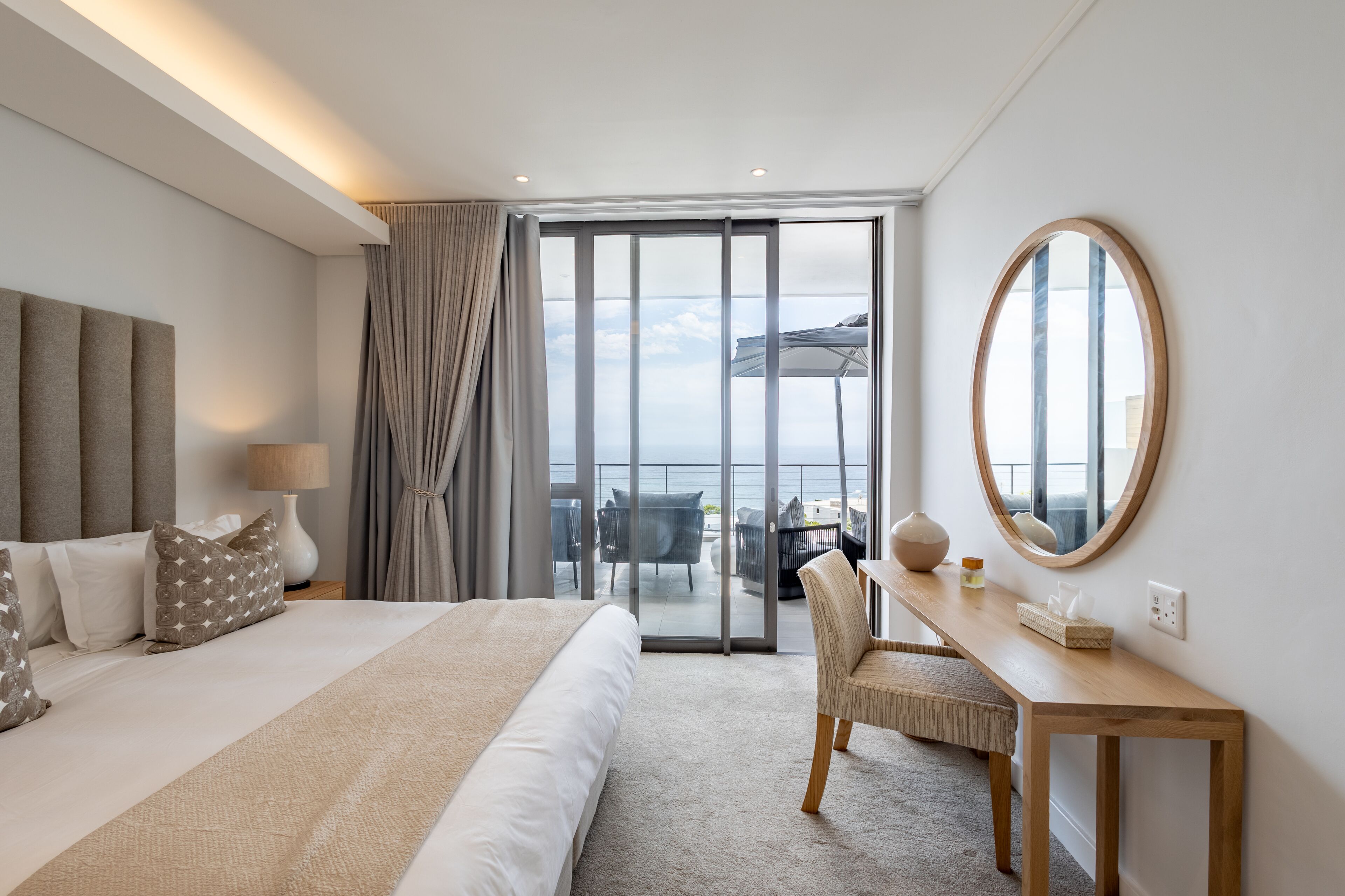 Habitación de hotel con una cama grande, un escritorio con silla, un espejo redondo y vista al mar a través de una puerta corredera de vidrio.