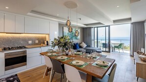 A modern kitchen with white cabinets, a wooden dining table set for six, and a large window overlooking the sea. - Plett Quarter Apartments (Plettenberg Bay)
