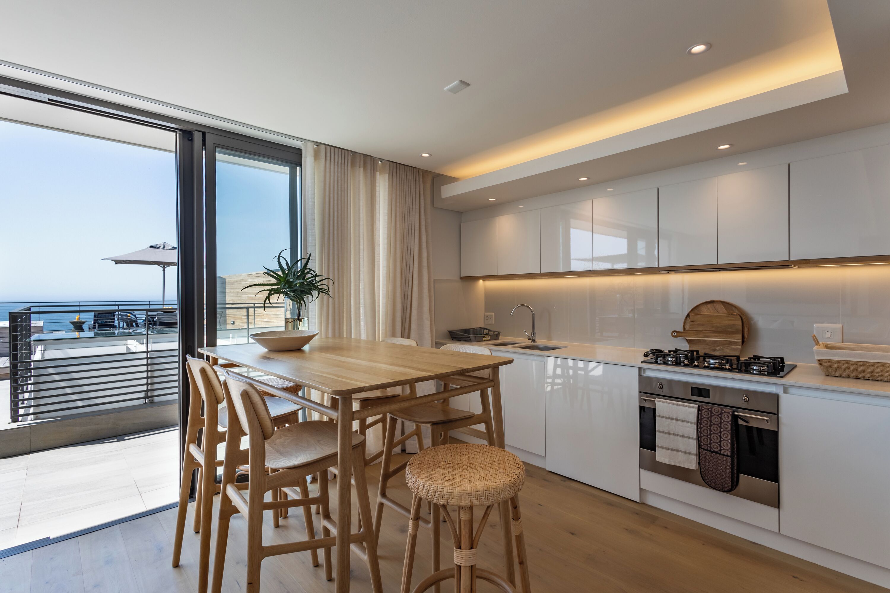 Una cocina moderna con una mesa y sillas de madera, un ventanal con vistas al mar y una pequeña isla con estufa.