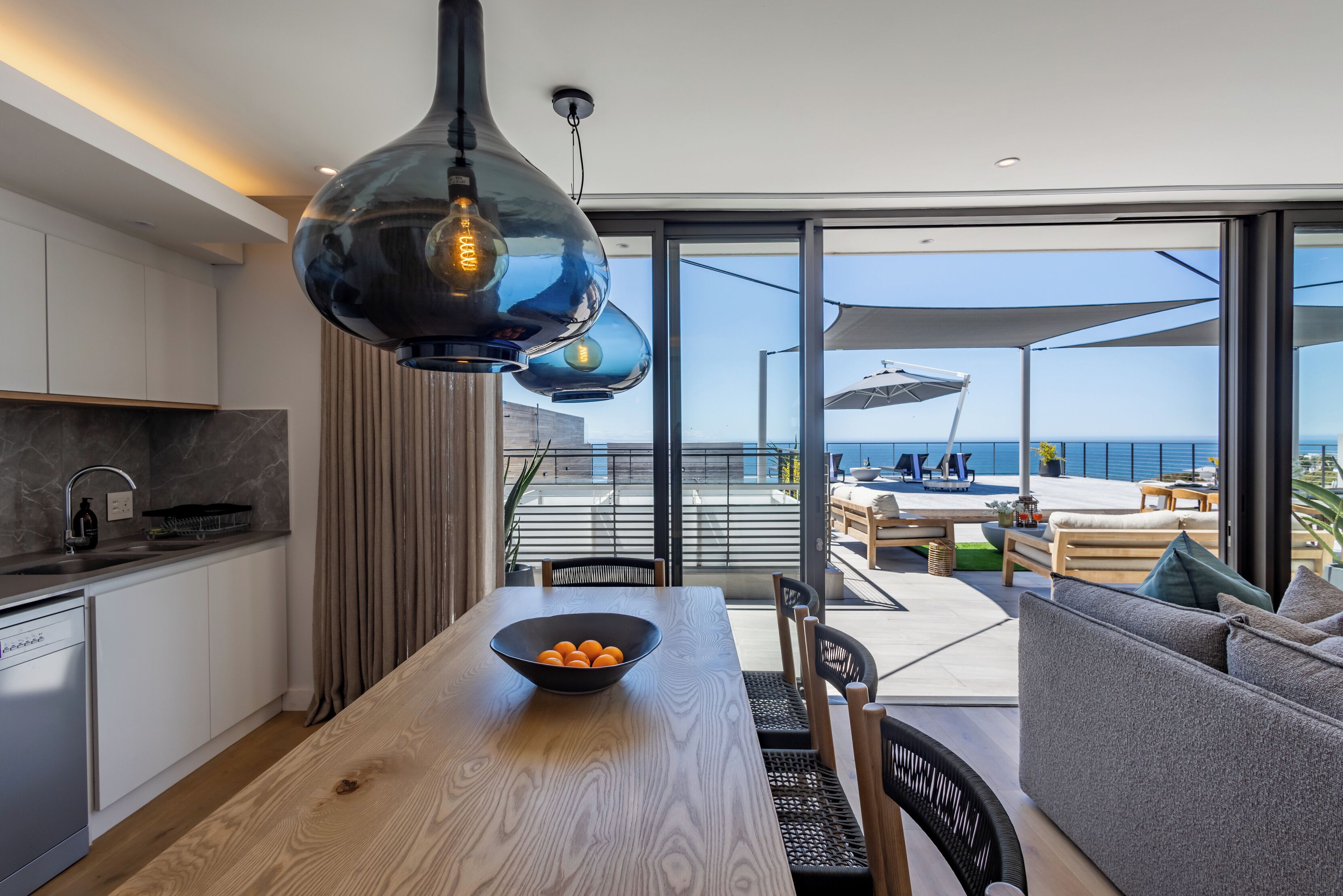 A modern kitchen with a wooden table, a bowl of oranges, and a view of the sea through large sliding doors.