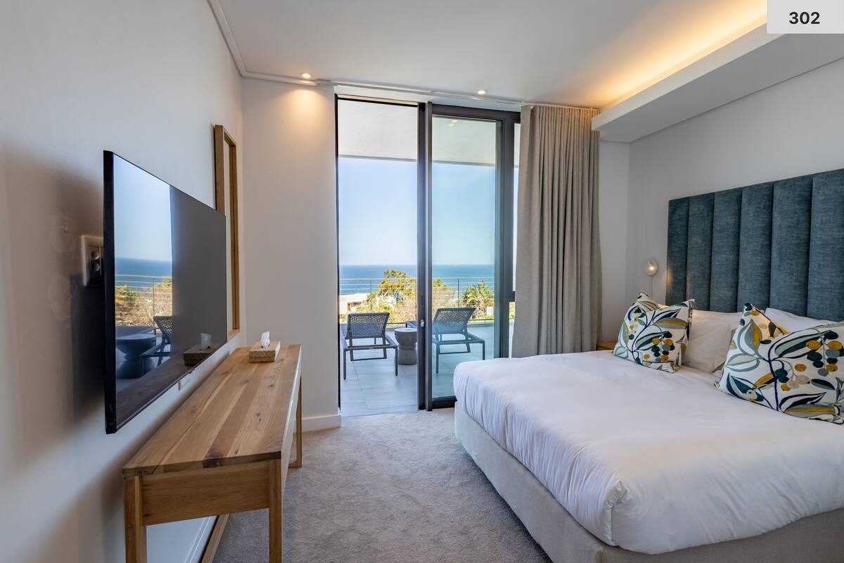 A modern hotel room with a large bed, a flat-screen TV, a wooden desk, and a balcony with a view of the sea.