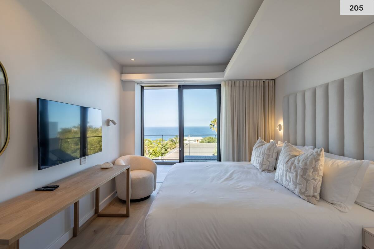 A modern hotel room with a large bed, a television on a wooden console, a round chair, and a balcony with a view of the sea.