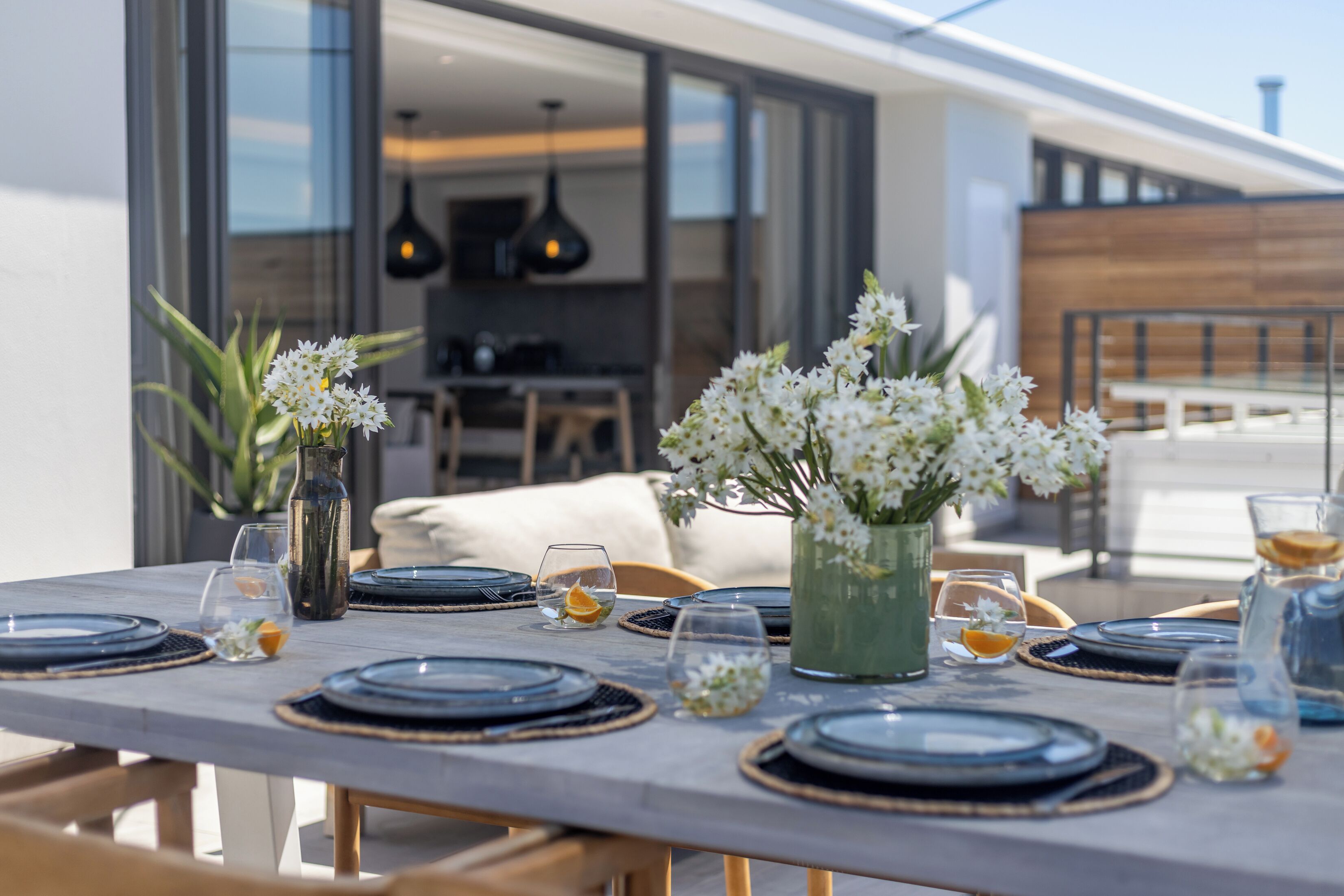 A dining table set with plates, glasses, and vases with flowers, overlooking a modern kitchen and dining area.