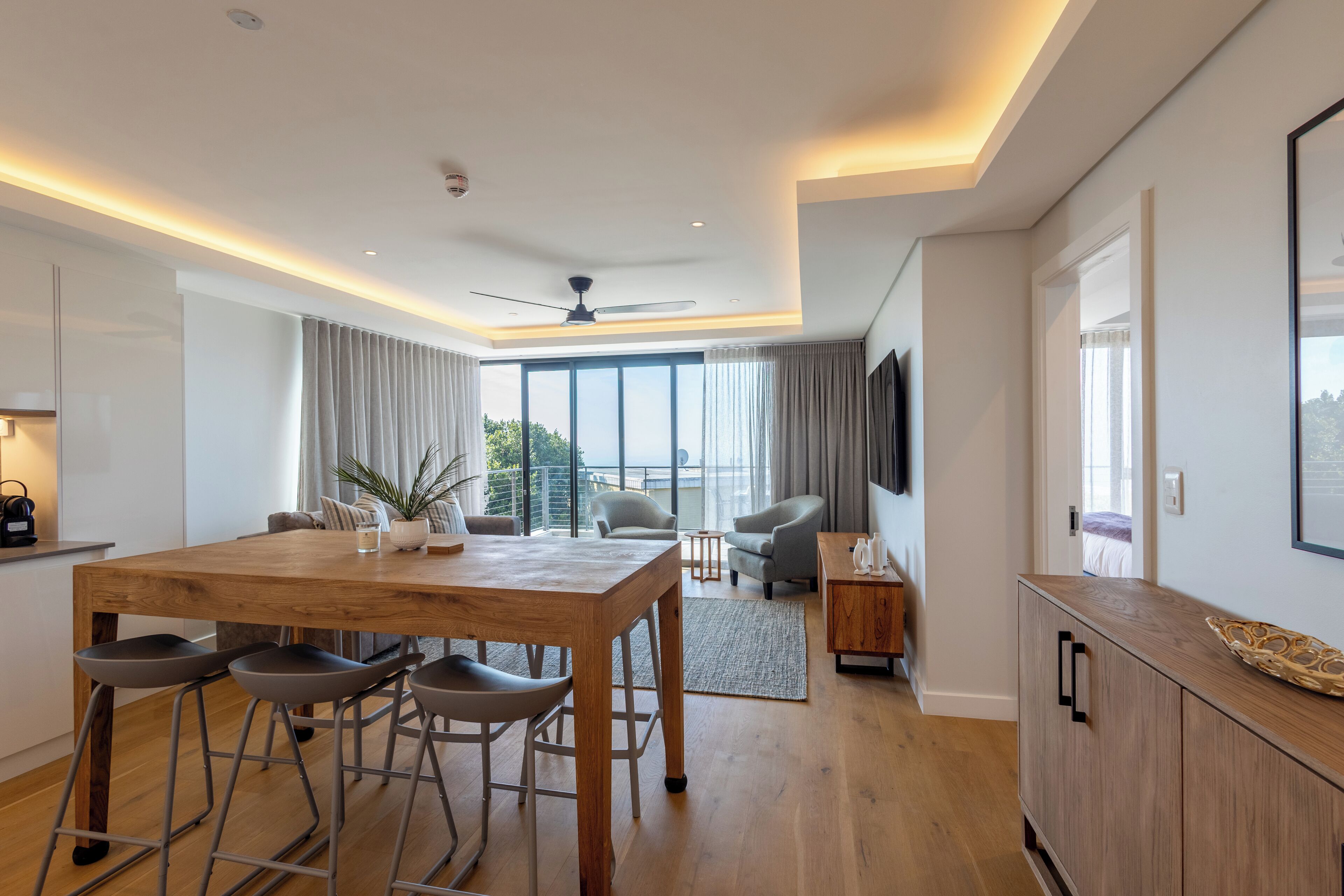 Een moderne woonkamer met een houten eettafel, barkrukken en een flatscreen tv aan de wand.