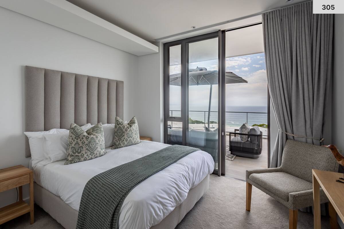 A modern hotel room with a large bed, a balcony with a view of the sea, and a chair.