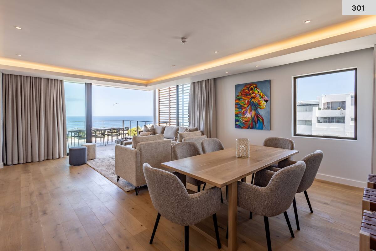 A modern living room with a wooden dining table and chairs, a sofa set, a large window with a view of the sea, and a colorful abstract painting on the wall.