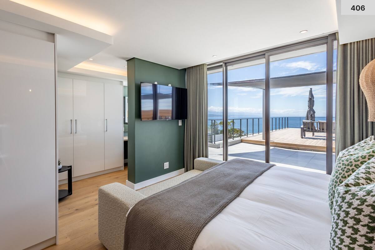 A modern hotel room with a large bed, a flat-screen TV, a balcony with a view of the sea, and a green accent wall.