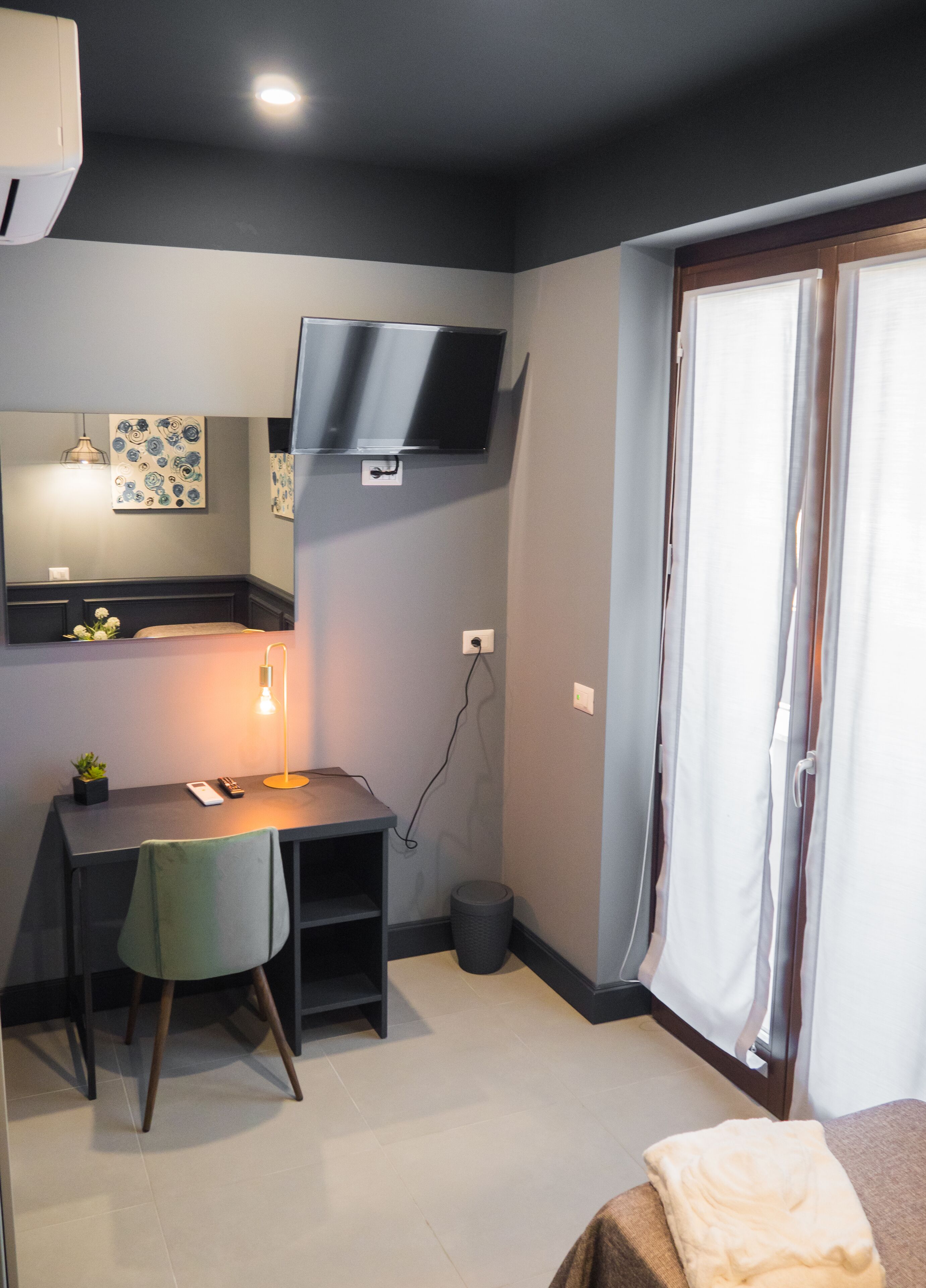A compact room with a desk, a chair, a bed, a TV mounted on the wall, and a window with curtains.