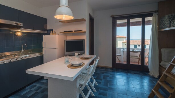 A compact kitchen with a central island, a built-in oven, and a refrigerator. There is a dining area with a table and chairs, and a large window offering a view of the outside. - Piazzetta Mono Blu Mare 50m From The Beac (Loiri Porto San Paolo)
