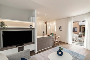 Uma sala de estar moderna com televisão de tela plana, sofá e mesa de centro. Há uma área de cozinha com mesa e cadeiras.