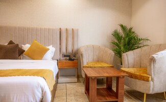 Una habitación de hotel con una cama, dos sillones, una mesita de madera, una mesita de noche con una lámpara y una planta en maceta.