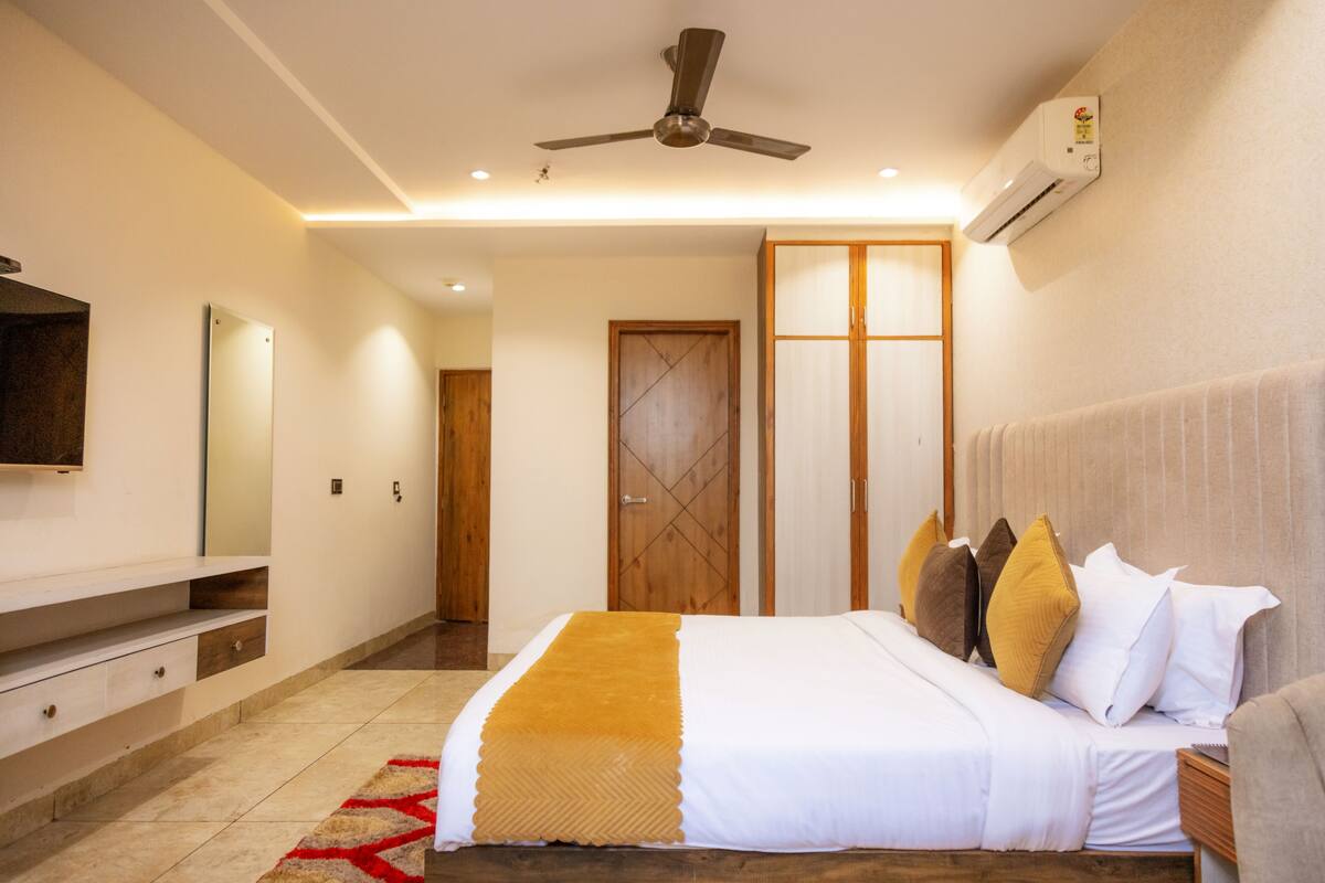 A modern hotel room with a large bed, a ceiling fan, an air conditioning unit, a wooden wardrobe, and a flat-screen TV mounted on the wall.