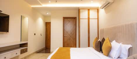 A modern hotel room with a large bed, a ceiling fan, an air conditioning unit, a wooden wardrobe, and a flat-screen TV mounted on the wall.