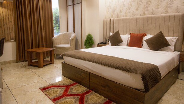 A bedroom with a large bed, a nightstand, a small table, a chair, and a window with curtains. - Hotel Eminence (Mohali)