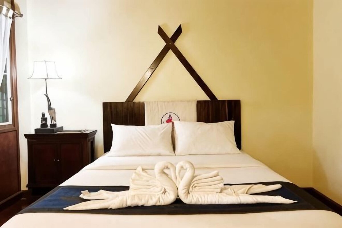 A hotel room with a bed, pillows, a headboard, a nightstand, a lamp, and towels arranged on the bed.