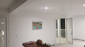 Une pièce avec un mur blanc, un tableau, un bureau avec une chaise et une porte donnant sur un balcon.