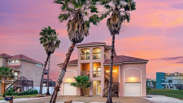 A multi-story residential building with a two-story garage, surrounded by palm trees, under a sunset sky. - Walk to the Beach | Golf Cart Rental, Beach Boardwalk, Balcony Views | AvantStay (Port Aransas)