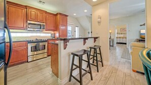 A kitchen with wooden cabinets, a microwave, stove, and a bar stool. - Walk to the Beach | Golf Cart Rental, Beach Boardwalk, Balcony Views | AvantStay (Port Aransas)