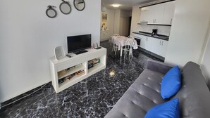 A modern living room with a flat-screen TV, a white entertainment unit with books, a grey sofa with blue cushions, and a dining area with a table and chairs. - Guayero 2 (Los Cristianos)