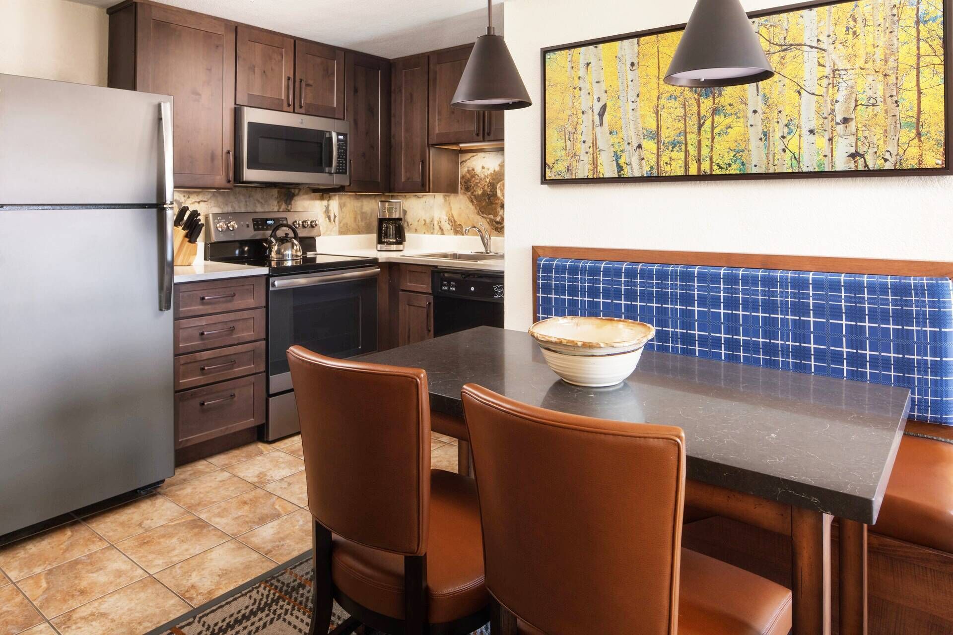 A kitchen with dark wooden cabinets, a stainless steel refrigerator, a microwave, a kettle, and a sink. There is a dining table with two chairs and a bowl on it.
