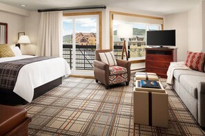 Ein Hotelzimmer mit Bett, einer Couch, einem Sessel, einem Fernseher und Blick auf Gebäude durch große Fenster.