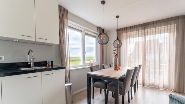 A modern kitchen with white cabinets, a black countertop, and a stainless steel sink. There is a dining table with chairs and a vase with flowers. The room has large windows with sheer curtains and a view of the outdoors. - Holiday Home near Scherpenisse Beach (Scherpenisse)