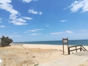 A beach with a wooden boardwalk leading to the water, a signpost, and beach umbrellas in the distance.