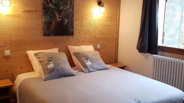 Ein Schlafzimmer mit Holz-Kopfteil, zwei Wandleuchten, einem großen Bett mit grauer Bettdecke, zwei Kissen mit Hirschmotiven und einem Fenster mit Vorhängen.