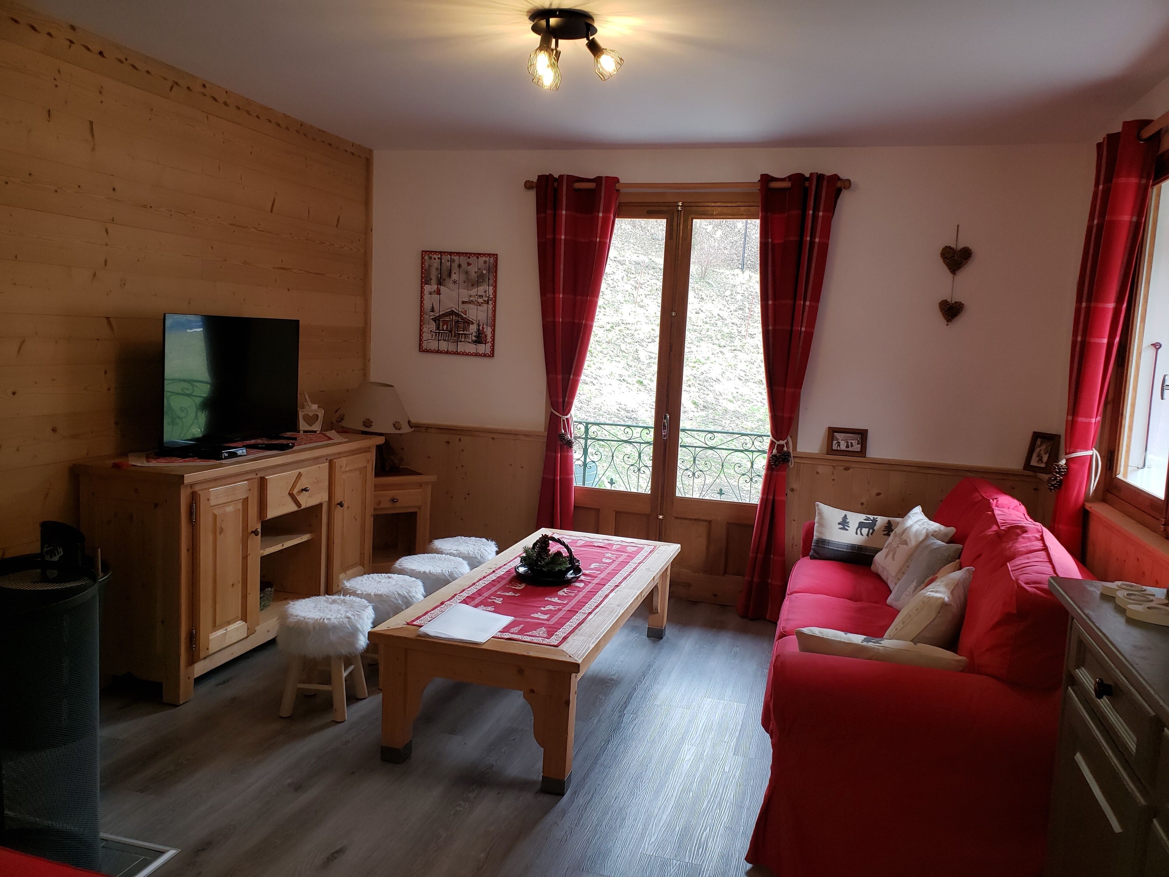 A cozy living room with a wooden floor, a television on a wooden cabinet, a red sofa, and a window with red curtains.