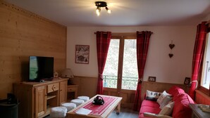Ein gemütliches Wohnzimmer mit Holzboden, einem Fernseher auf einer Holzkommode, einem roten Sofa und einem Fenster mit roten Vorhängen.