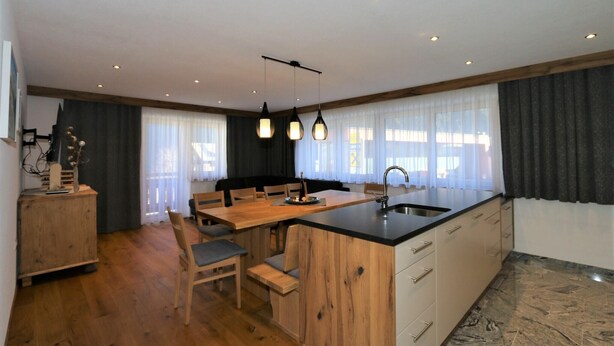 A modern kitchen with a wooden island, a dining area with chairs, and a large window with curtains. - Beautiful holiday flat in Krimml near waterfalls (Krimml)