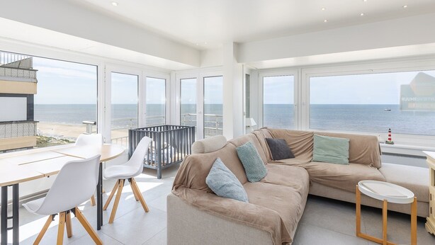 Een ruime woonkamer met een groot raam met uitzicht op de zee, een bank, een eettafel en een klein bijzettafeltje.