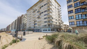 Een meerlaags gebouw met balkons, een zandig gebied ervoor en een heldere lucht.