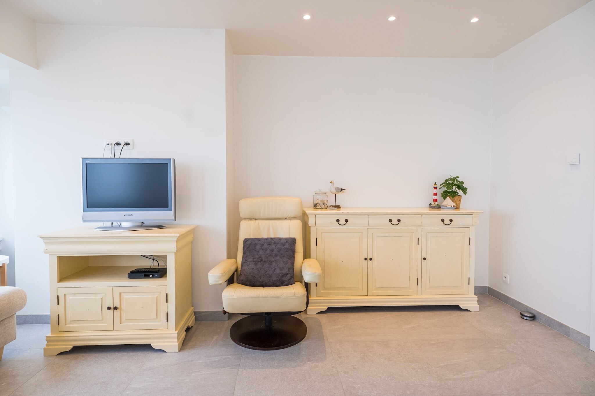 A living room with a television on a stand, a cream-colored armchair, a cabinet with drawers, and a small plant on top.
