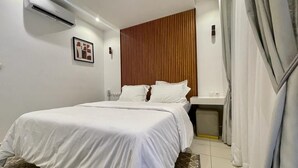 A bedroom with a bed, a wooden panel wall, an air conditioning unit, a bedside table, and a trash bin. - Swanky  Apartments (Douala)