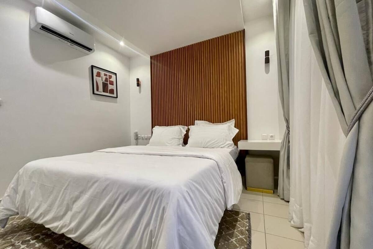 A bedroom with a bed, a wooden panel wall, an air conditioning unit, a bedside table, and a trash bin.