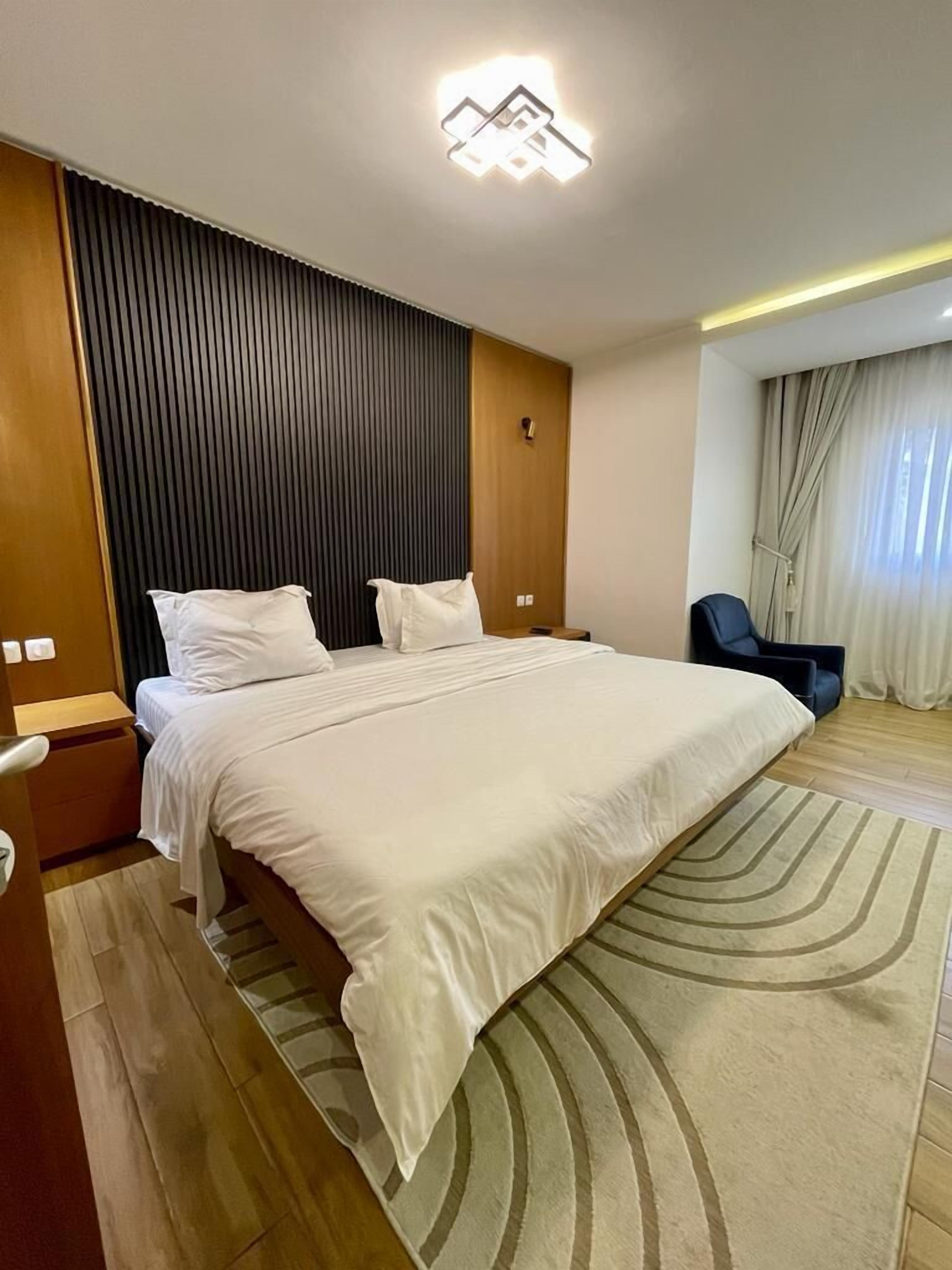 A modern hotel room with a large bed, wooden panel wall, and a view of the outdoors through a window.