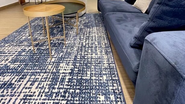 A modern living room with a blue sofa, a round coffee table, and a blue patterned rug. There is a kitchen in the background with white cabinets and a dining area with two blue chairs. - Swanky  Apartments (Douala)