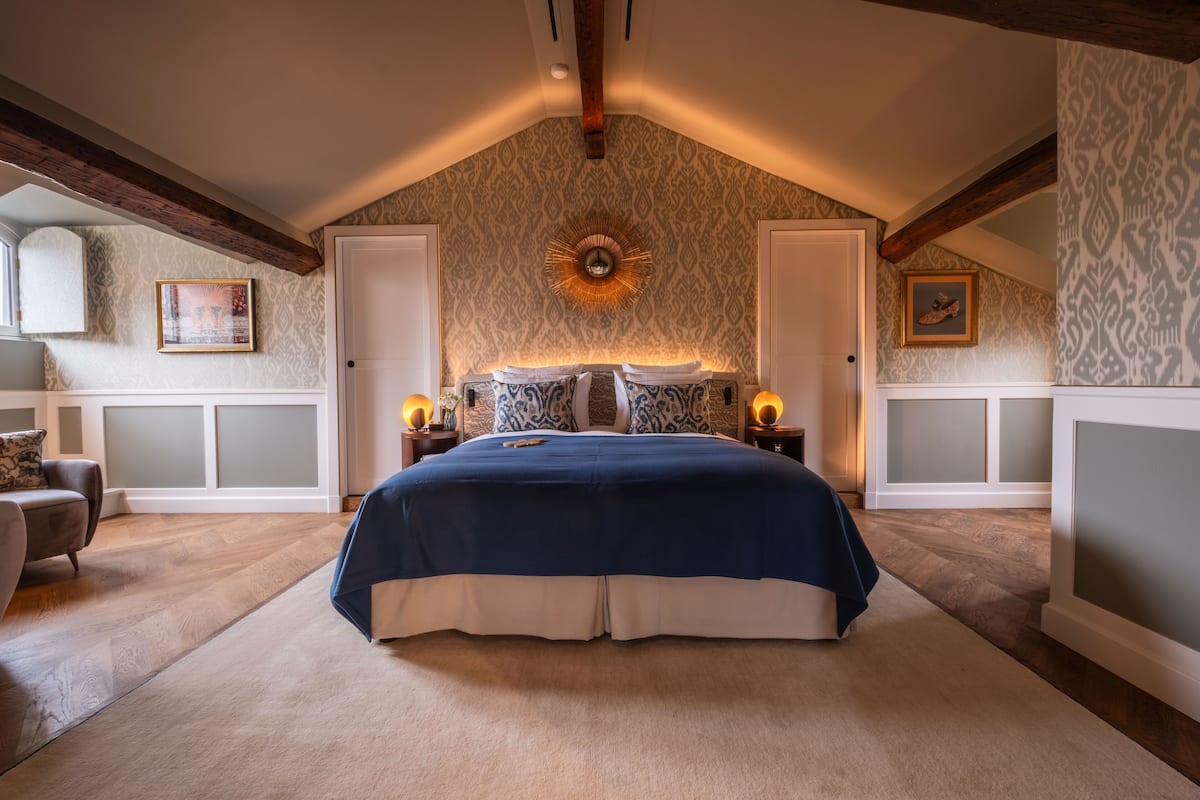Een slaapkamer met een groot bed, houten balken, behang met een patroon en twee bedlampen.