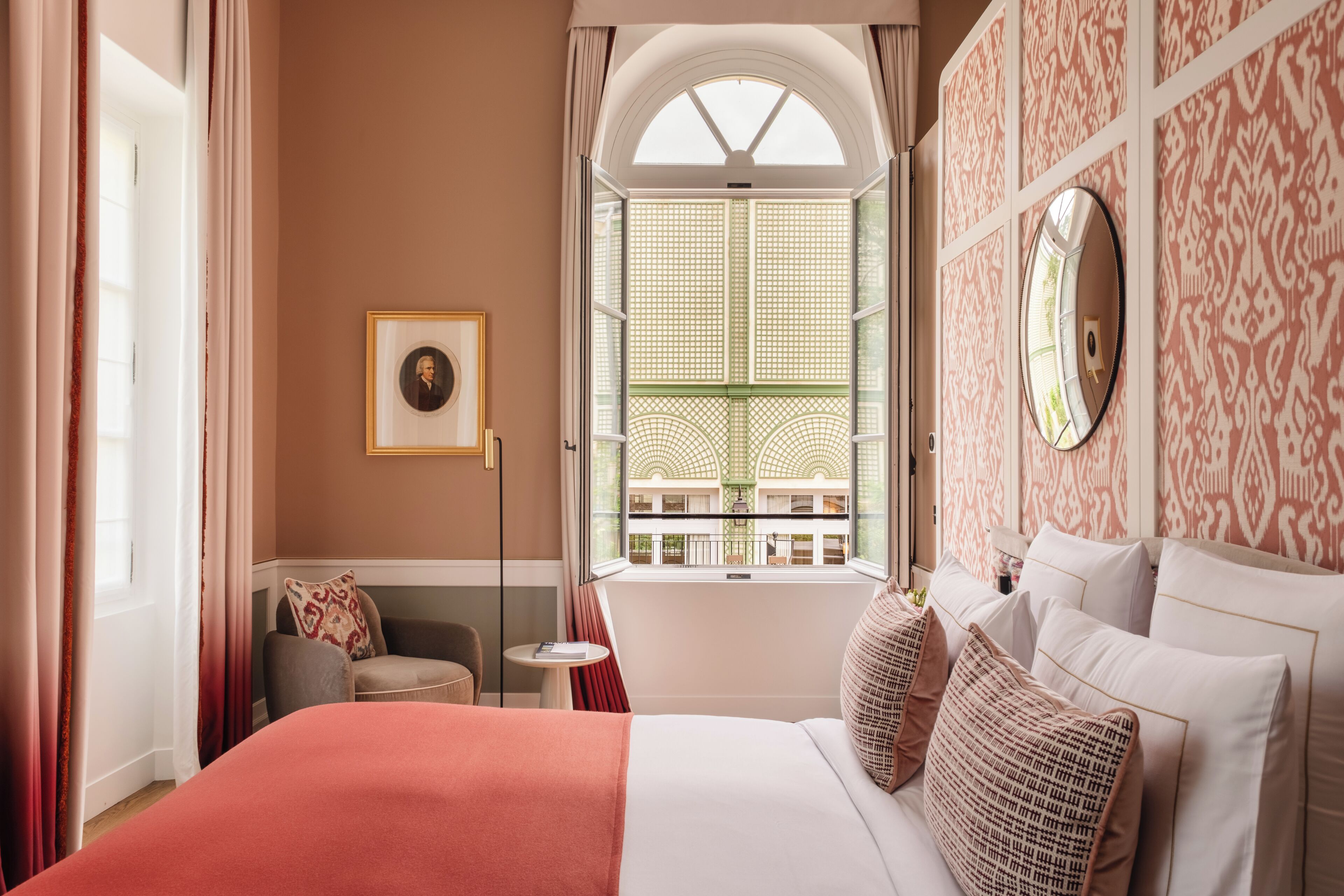 Une chambre d’hôtel avec un grand lit, une petite table, une chaise, une fenêtre donnant sur un immeuble et un tableau encadré au mur.