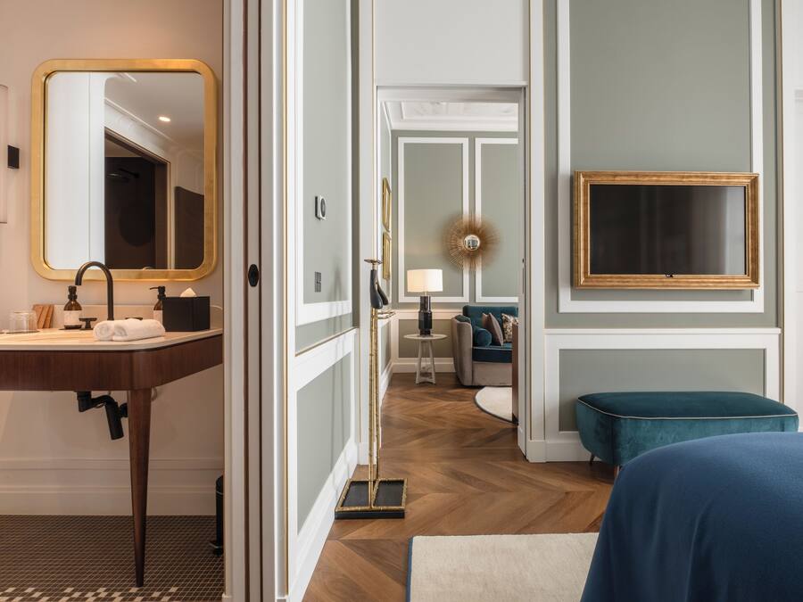 A hotel room with a wooden vanity, a large mirror, a bed with a blue bedspread, a teal ottoman, and a wall-mounted TV.
