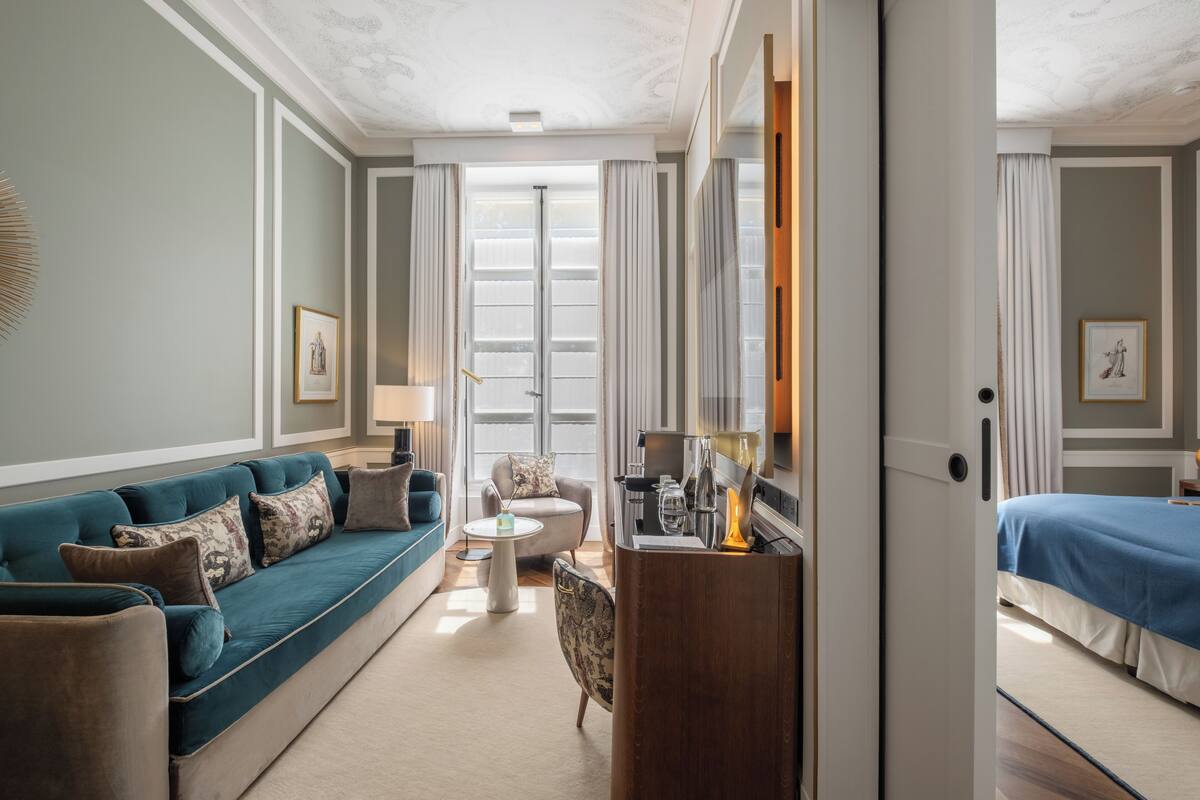 Een hotelkamer met een blauwgroene bank, een klein rond tafeltje, een houten dressoir en een bed met een blauwe spreidekking.