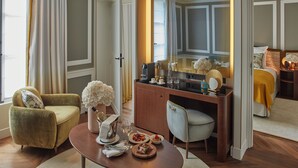 A well-lit room with a dining table set for two, a sofa, a desk with a mirror, and a bed with a yellow blanket. - Hôtel Les Lumières - Relais & Châteaux  (Versailles)
