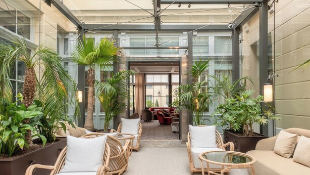A spacious indoor atrium with wicker furniture, potted plants, and a glass ceiling. - Hôtel Les Lumières - Relais & Châteaux  (Versailles)