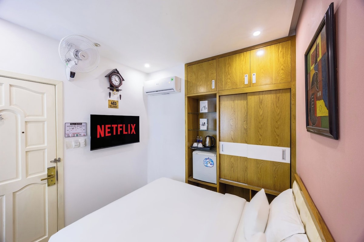 A hotel room with a bed, a wall-mounted TV, a clock, a fan, and a wardrobe.
